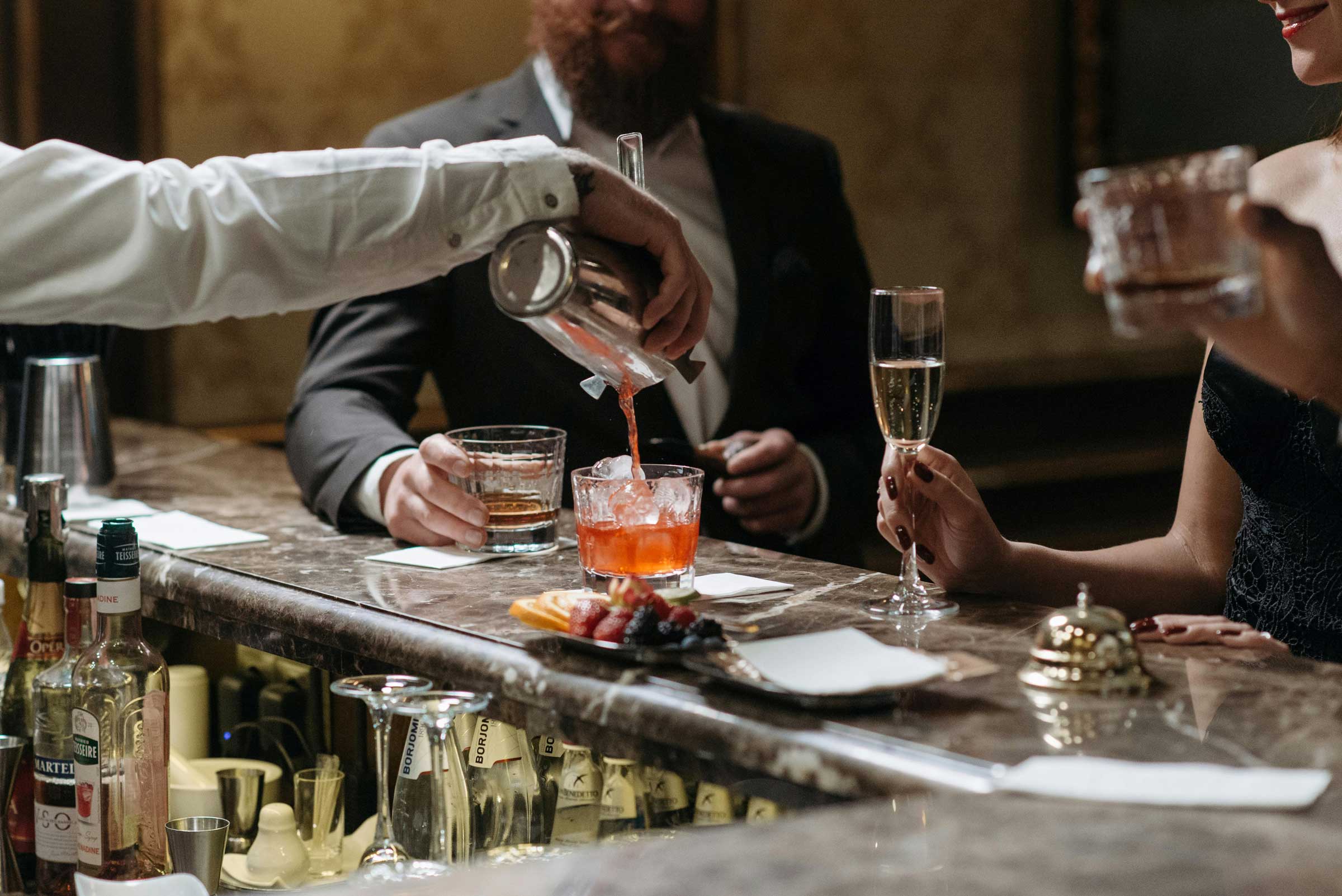 Man in a suit enjoys a cocktail while the bartender makes another on the bar.